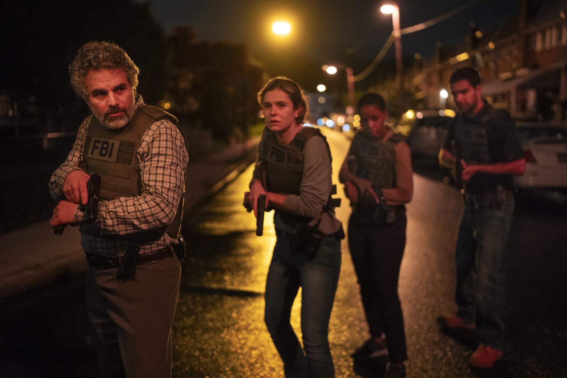 A white middle aged man wearing an FBI vest stands in the street at night, gun drawn, with three young people hovering behind him in similar stances.