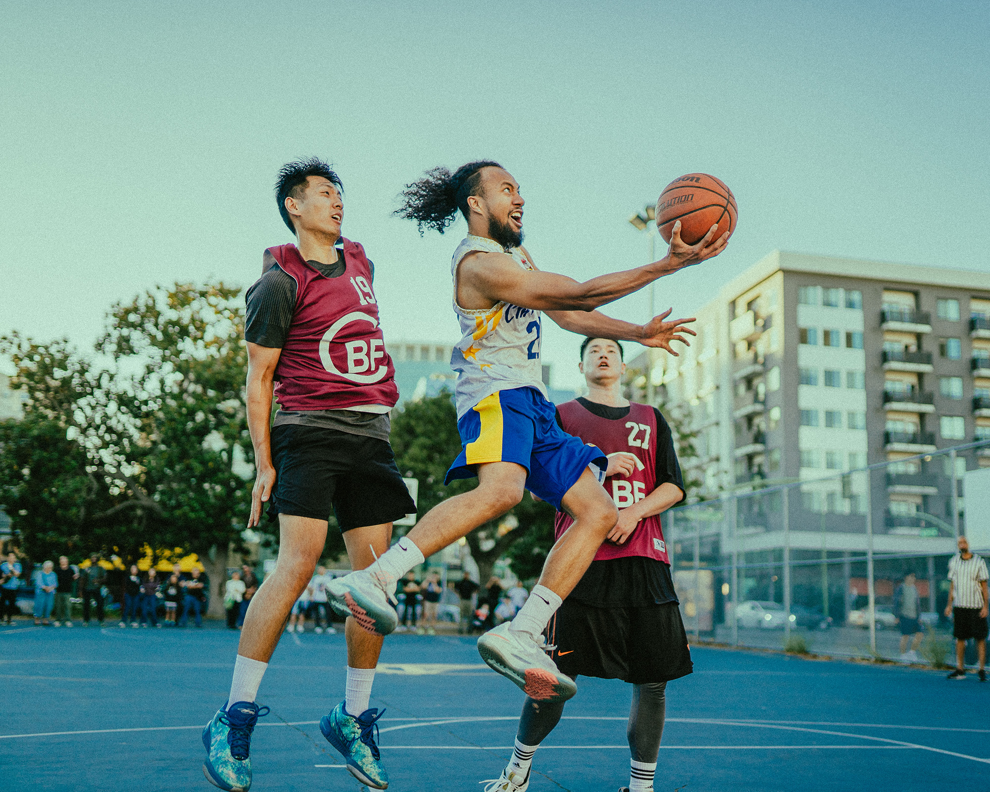 On an outdoor basketball court, a Filipino American player scoops the ball toward the basket as two opponents look on.