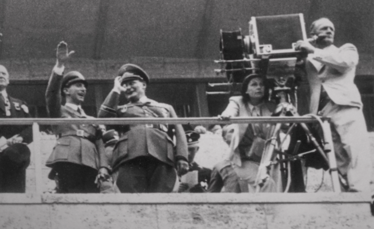 Two smiling men in nazi uniforms stand on a platform saluting at the crowd and smiling. Next to them is a woman seated behind a camera with another man at her side.