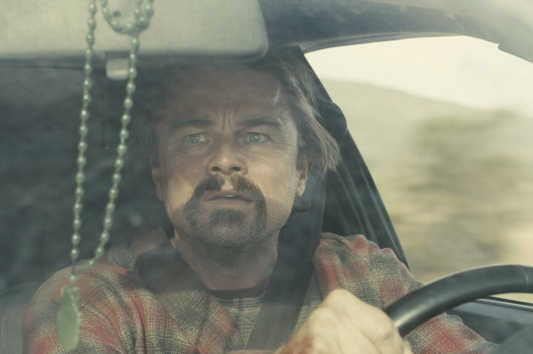 A scruffy middle-aged white man wearing a flannel shirt sits at a driver's seat and glances in his rearview mirror, a scared expression on his face.