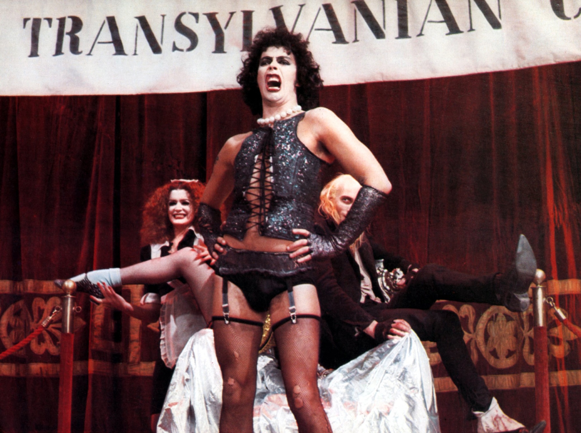 A white man in garish make-up wearing a corset, stockings and garter belt performs on a stage with a strange woman and man posing behind him.