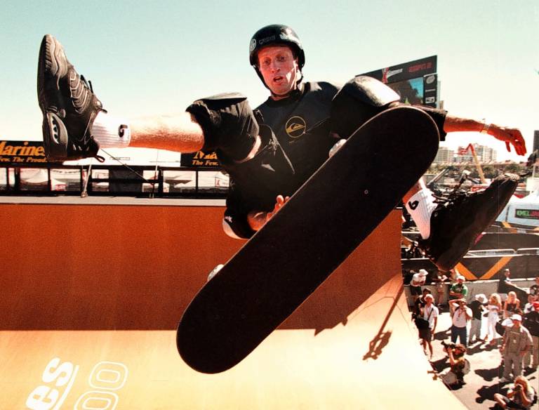 A white man in mid-air at the top of a half-pipe kicks out one leg from his skateboard.