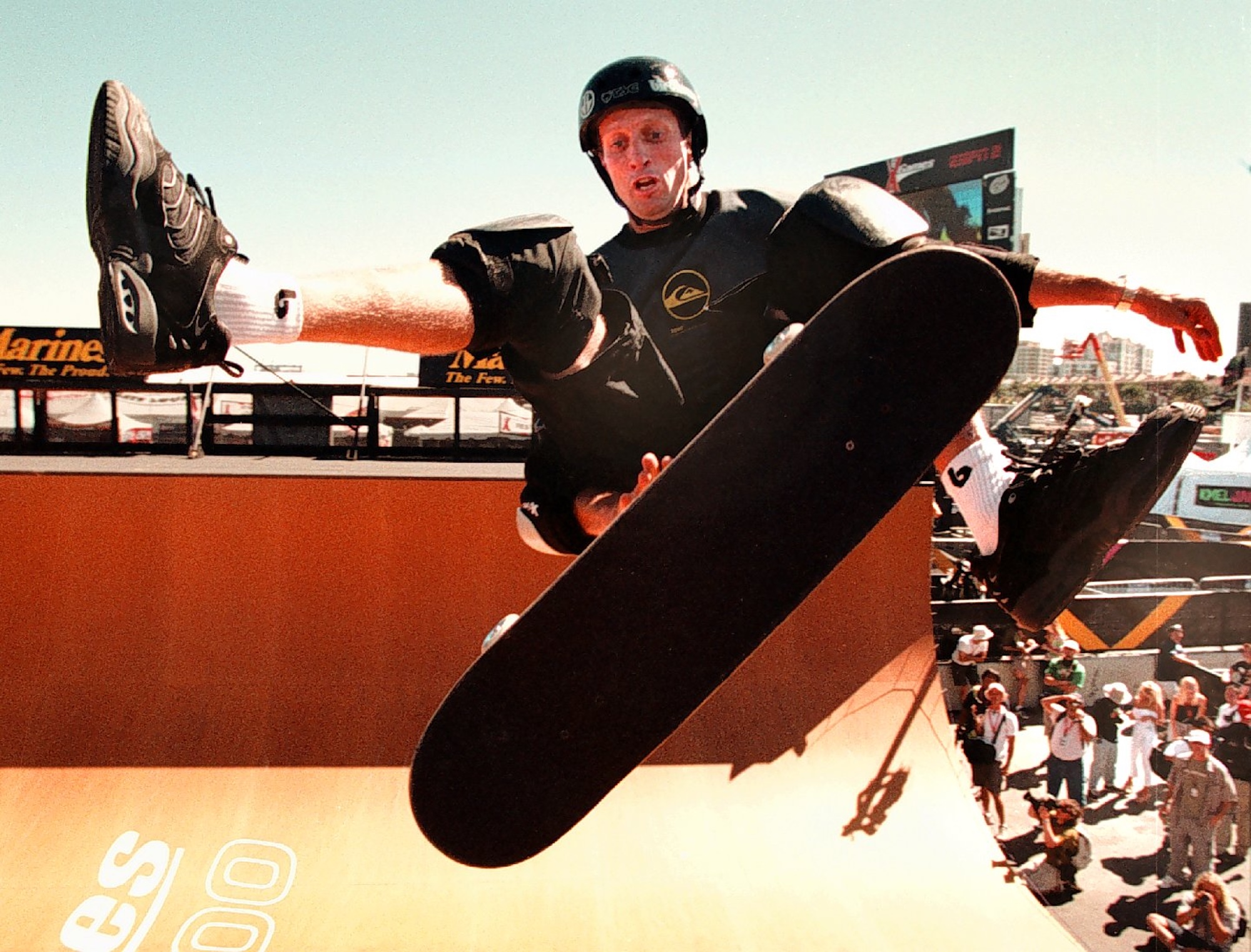 A white man in mid-air at the top of a half-pipe kicks out one leg from his skateboard.