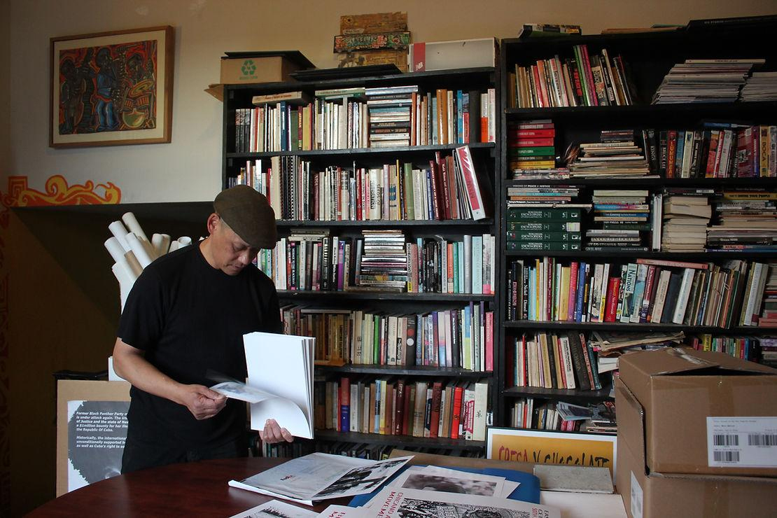 A man standing, reading a book, while standing in front of a bookcase. 