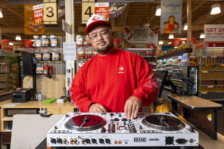 A DJ poses for a portrait at his turntable, which is set up inside a grocery store.
