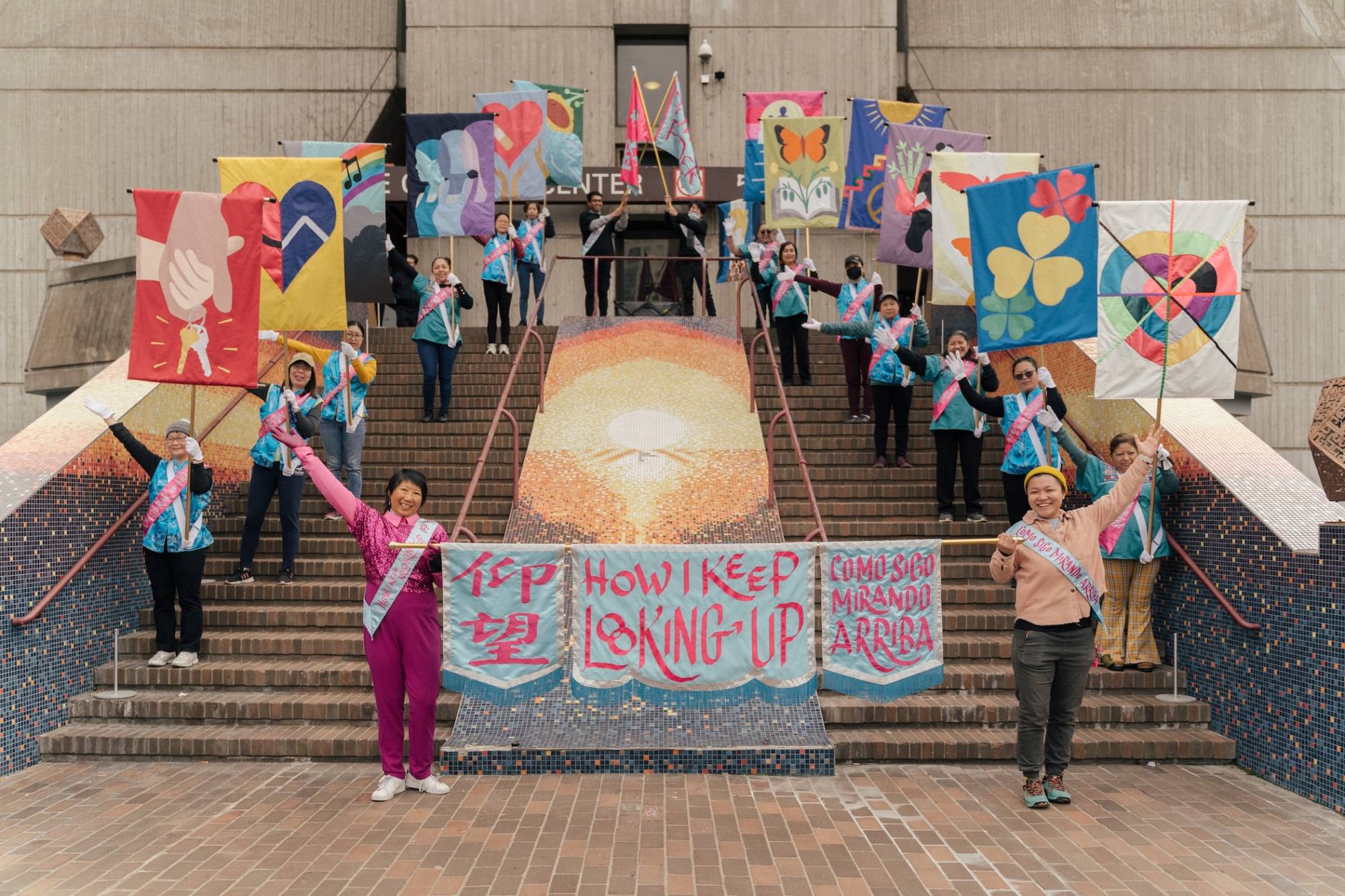 about 15 people stand on the steps of a cultural center holding flags and a sign that reads 'how I keep looking up'