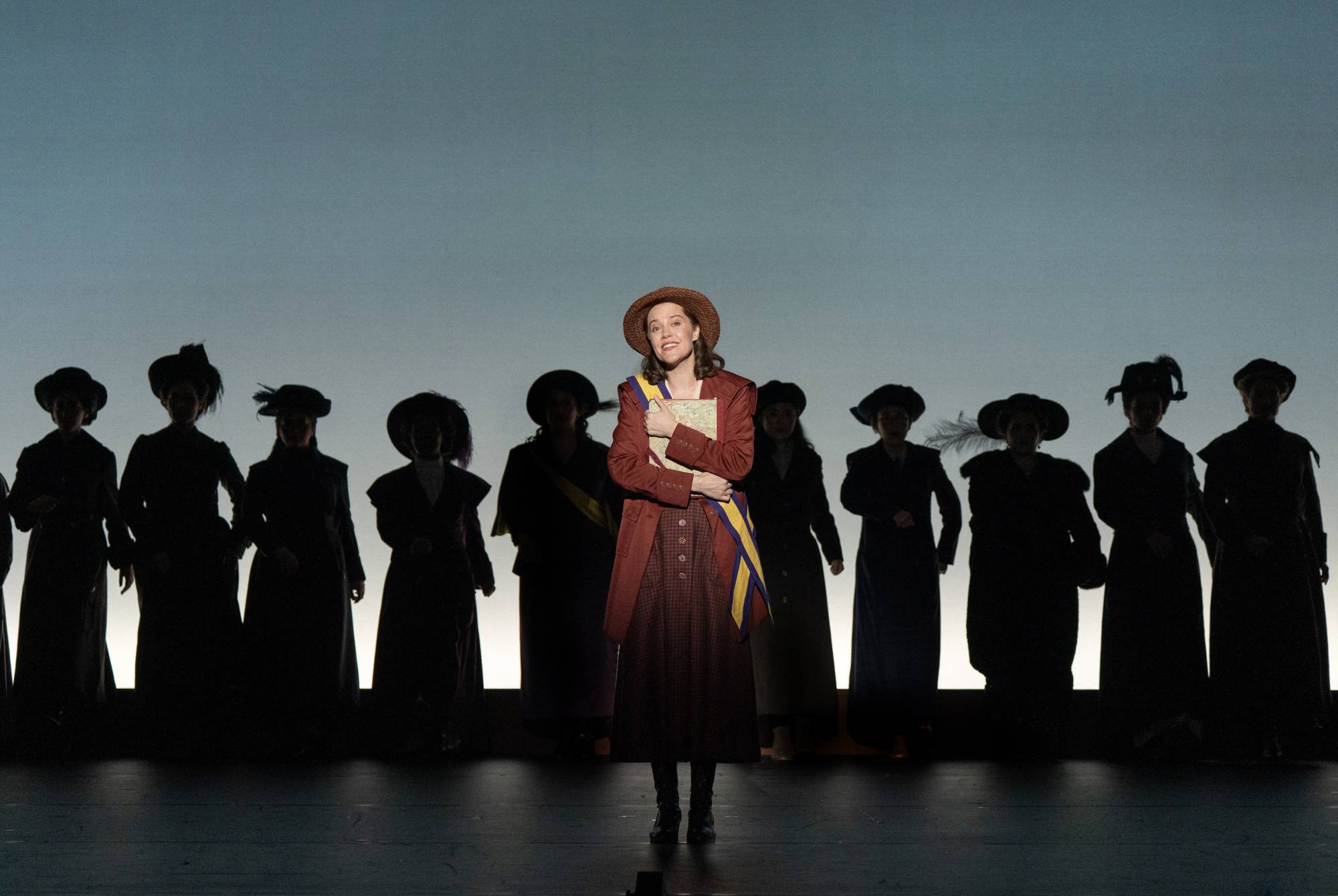 a woman stands on stage in front of other women in a play about the suffragettes