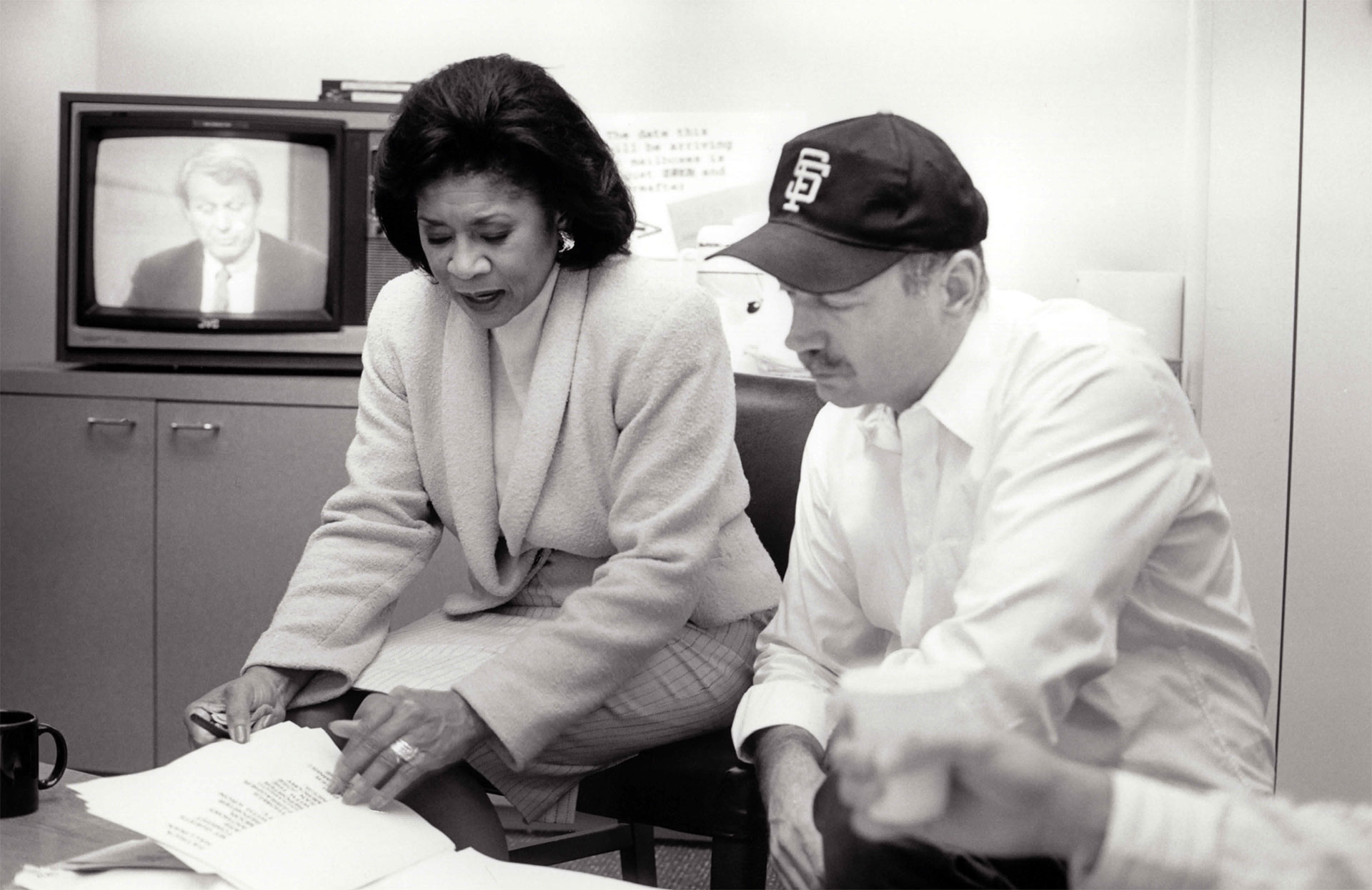 A Black woman and white man having a discussion looking at prepared sheets of paper with an old-style CRT television in the background