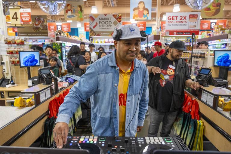 A DJ in a blue denim shirt works the turntable inside a crowded grocery store.