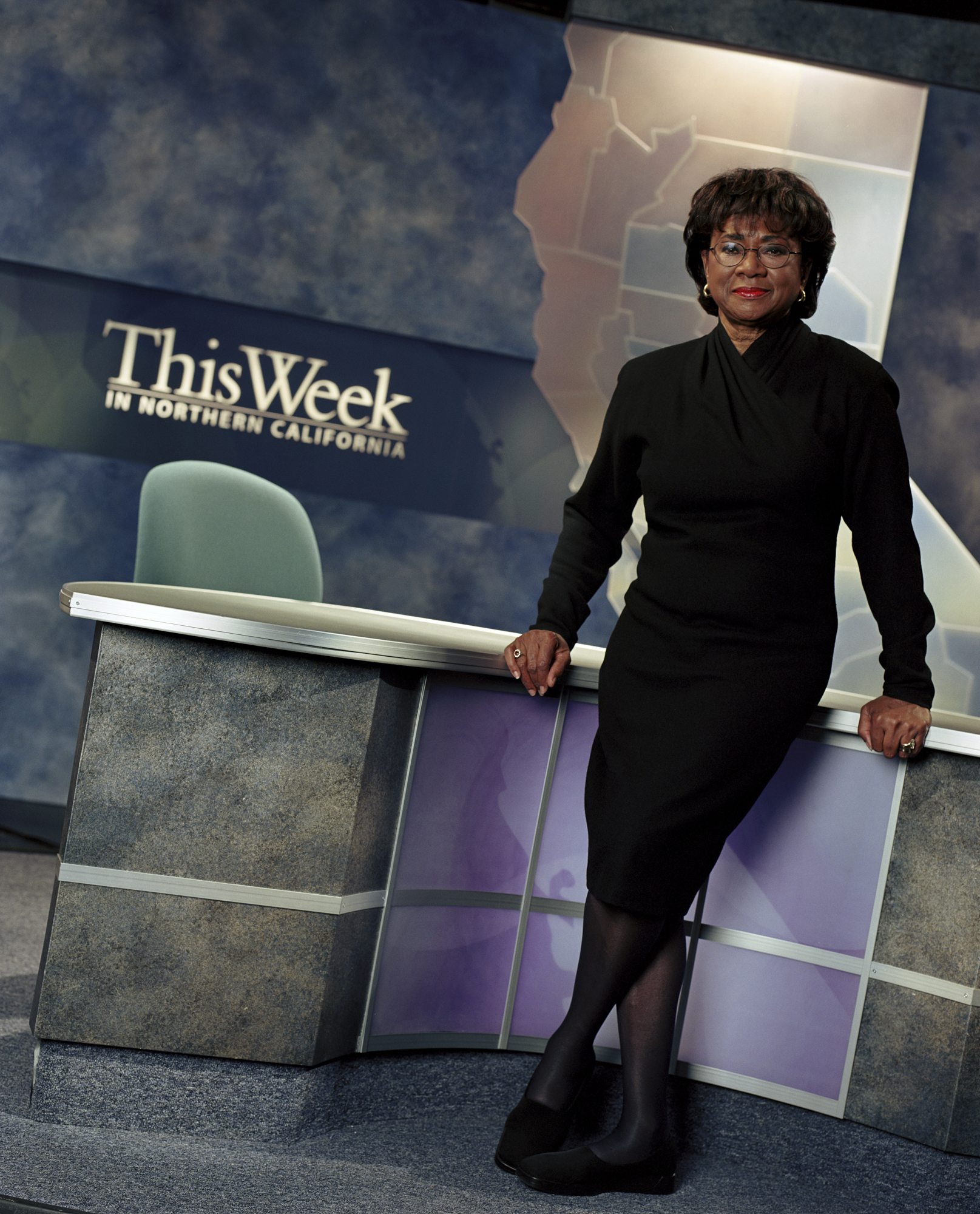 A person leans on a desk in a TV studio with the words "This Week in Northern California" written on the wall behind her.