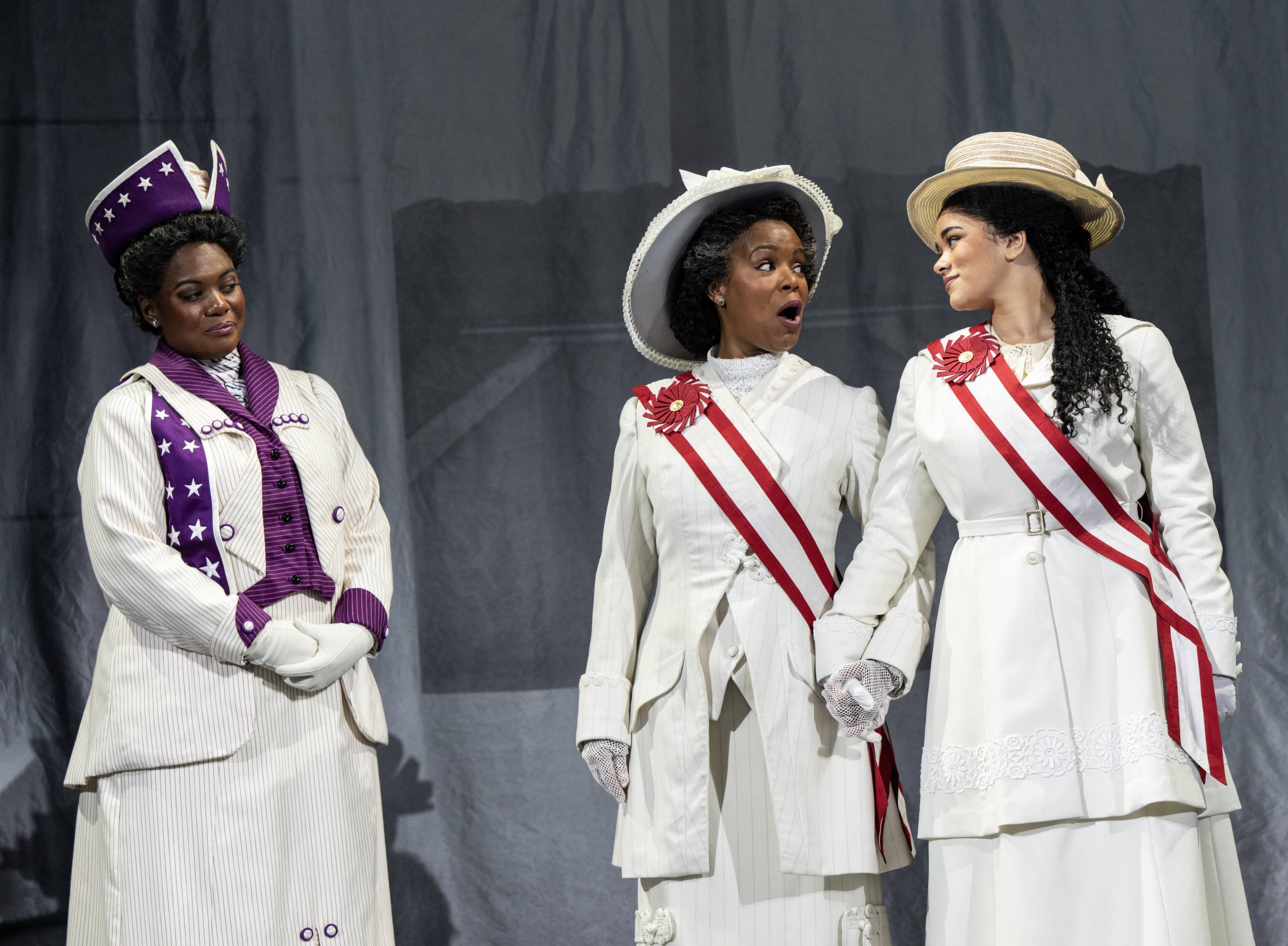 three women stand on stage in suffagette clothing in a play