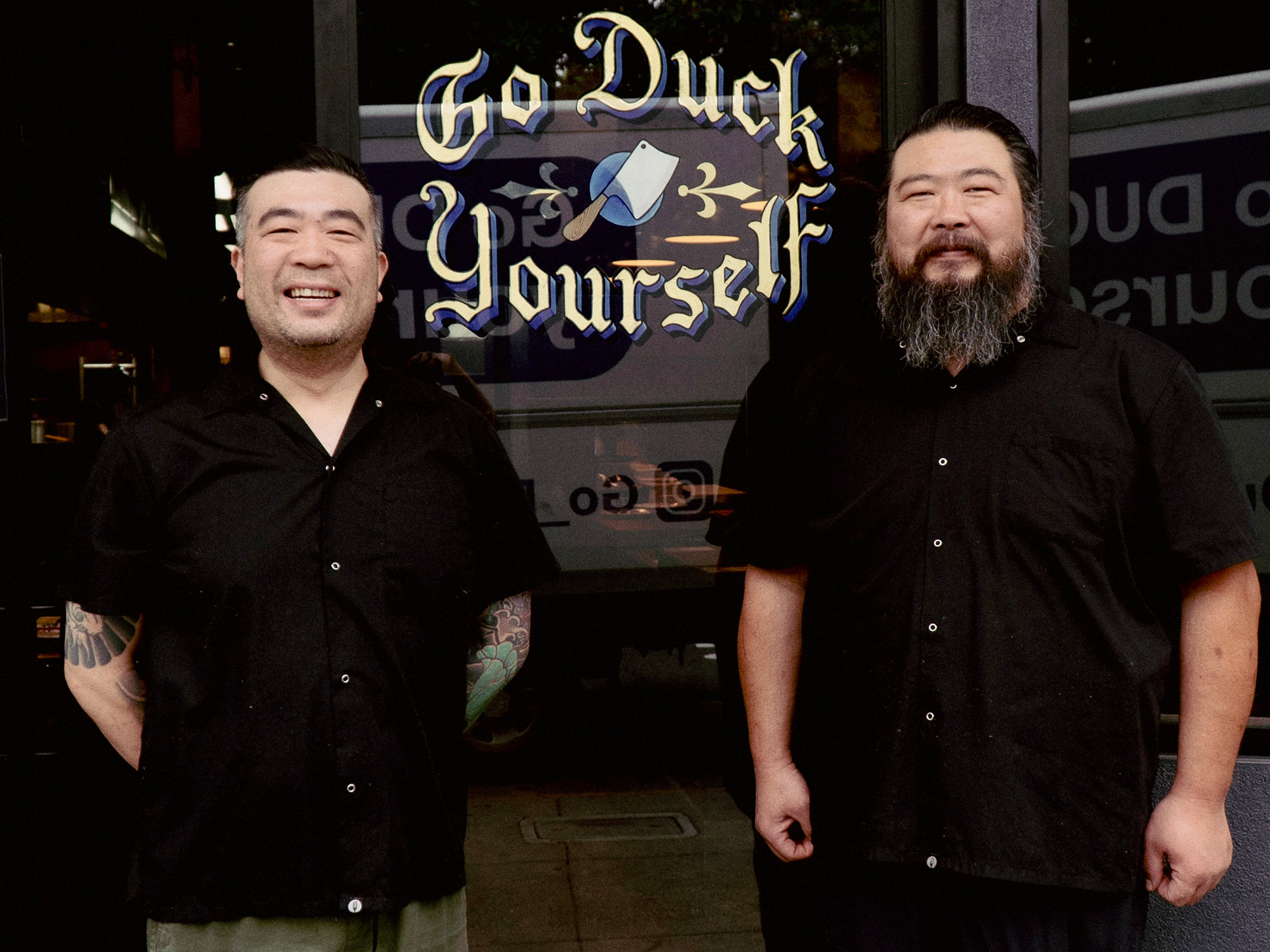 Two Asian men in black stand in front of a restaurant. The sign in the window reads, "Go Duck Yourself."