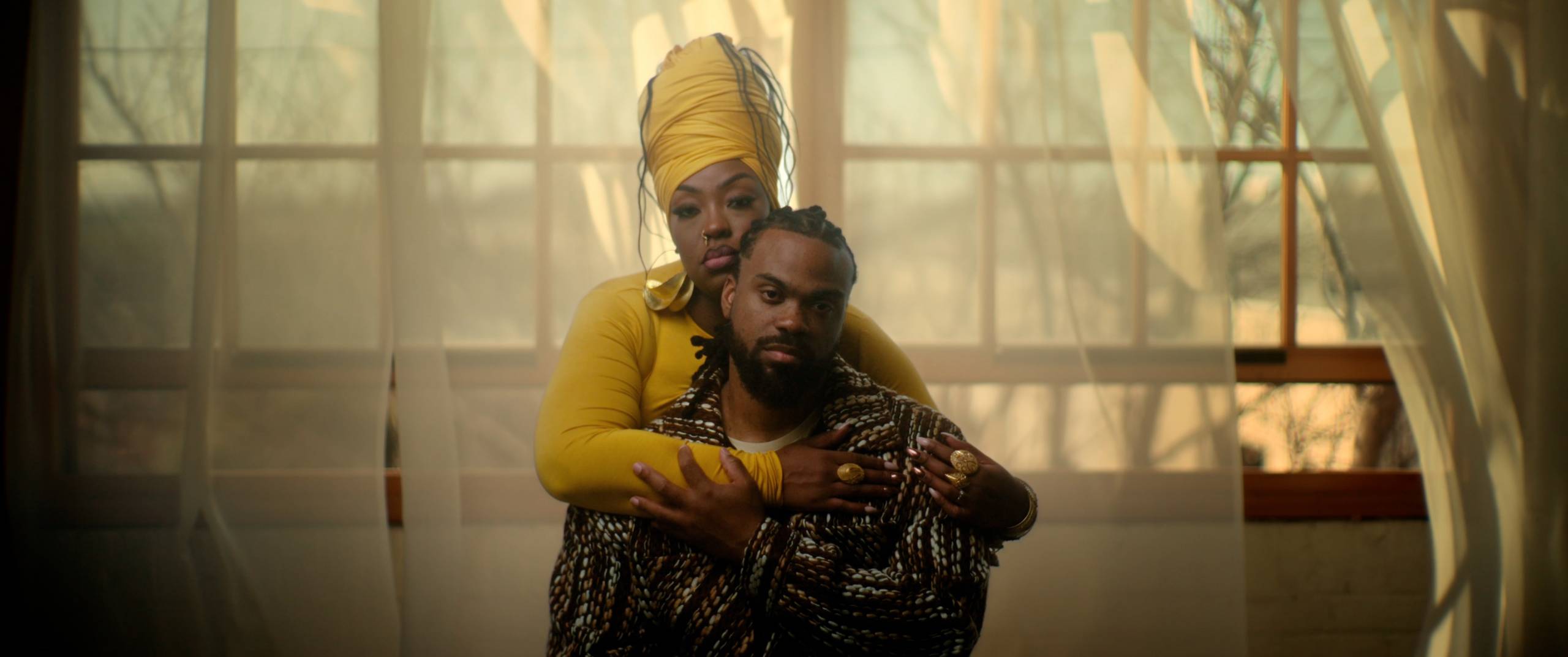 A woman wearing a yellow shirt and hair wrap stands behind a hand, arms around his shoulders in an embrace, as they pose for a photo.