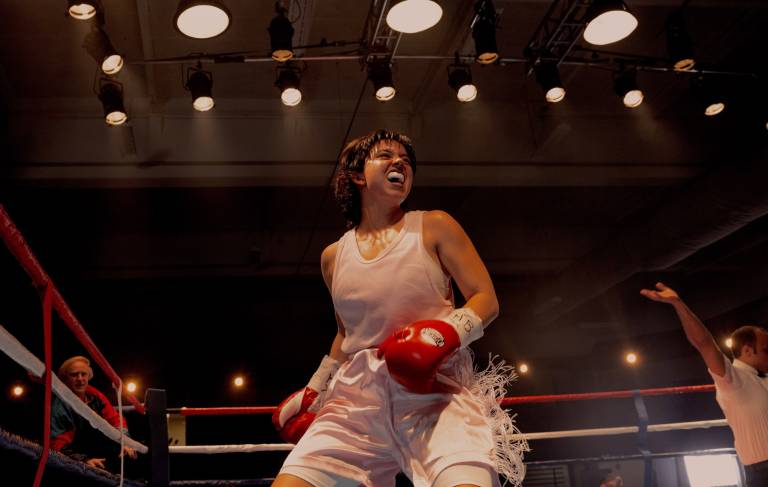 A white woman snarls and sweats inside a boxing ring triumphantly, wearing pink and white tank and shorts, and red boxing gloves.