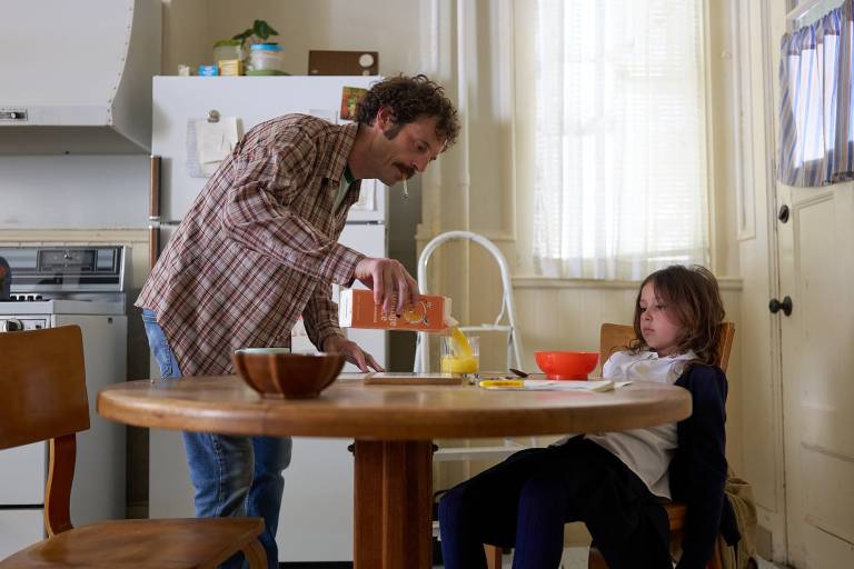 An average kitchen interior. A young white girl sits slumped at a round kitchen table, while her dad, wearing a flannel shirt and blue jeans, cigarette in mouth, pours her an orange juice.
