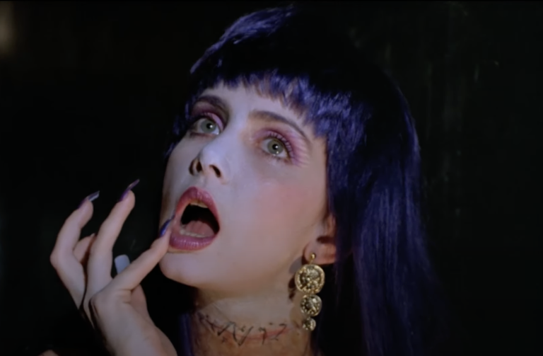 A pale beautiful woman with purple hair and a dead stare creepily touches her open lips with her little finger.