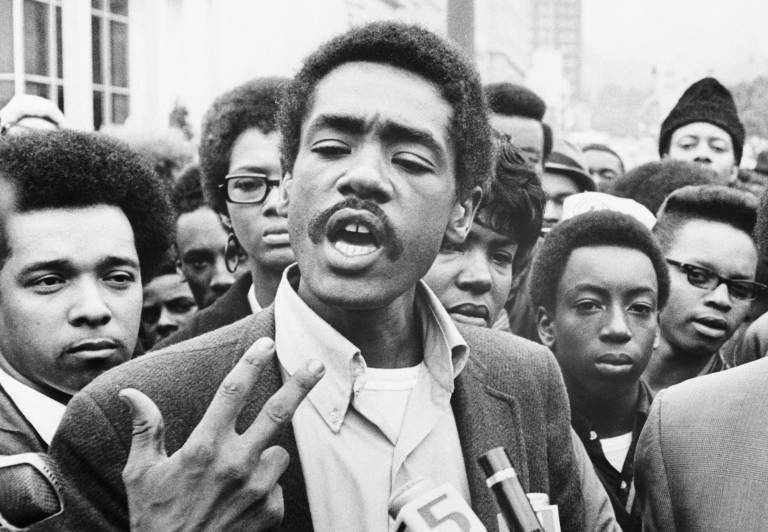 A 31-year-old Black man in a collared shirt and suit jacket speaks and gestures with his right hand, surrounded by a crowd of young Black supporters