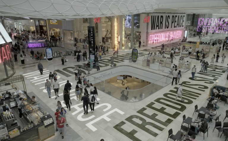 A busy shopping mall interior, viewed from above. Projections of phrases from George Orwell’s ‘1984’ are projected onto the floors and walls while shoppers remain oblivious. The projections read 'ignorance is strength,' 'war is peace,' and 'freedom is slavery.'