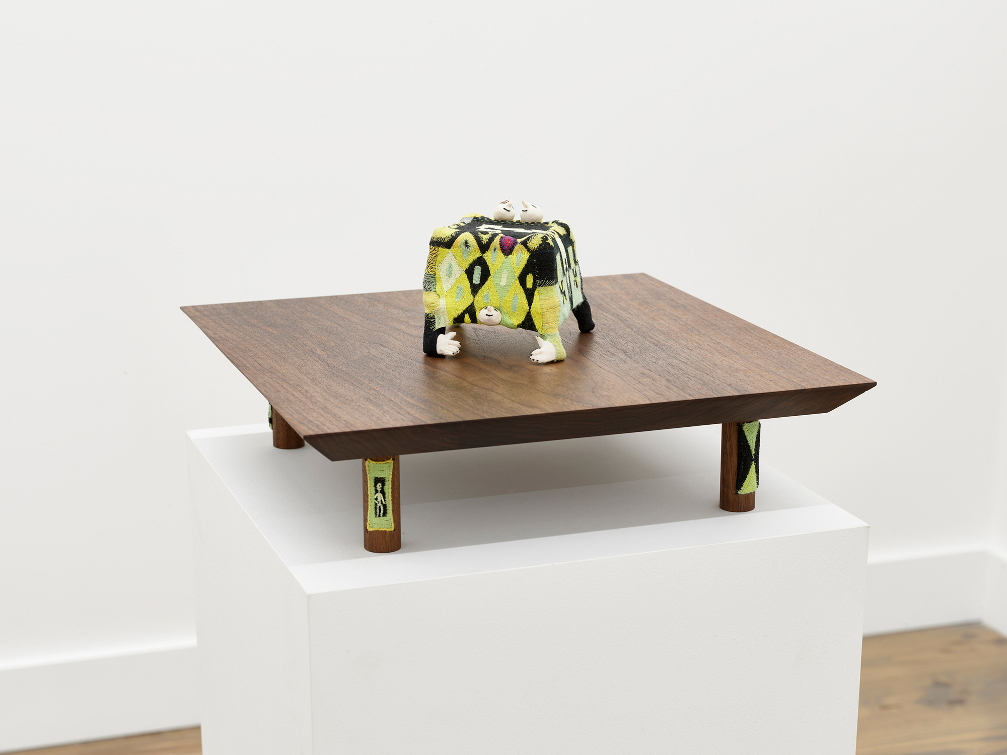 woven-covered sculpture of two heads in bed with head and arms underneath, on wooden pedestal with feet