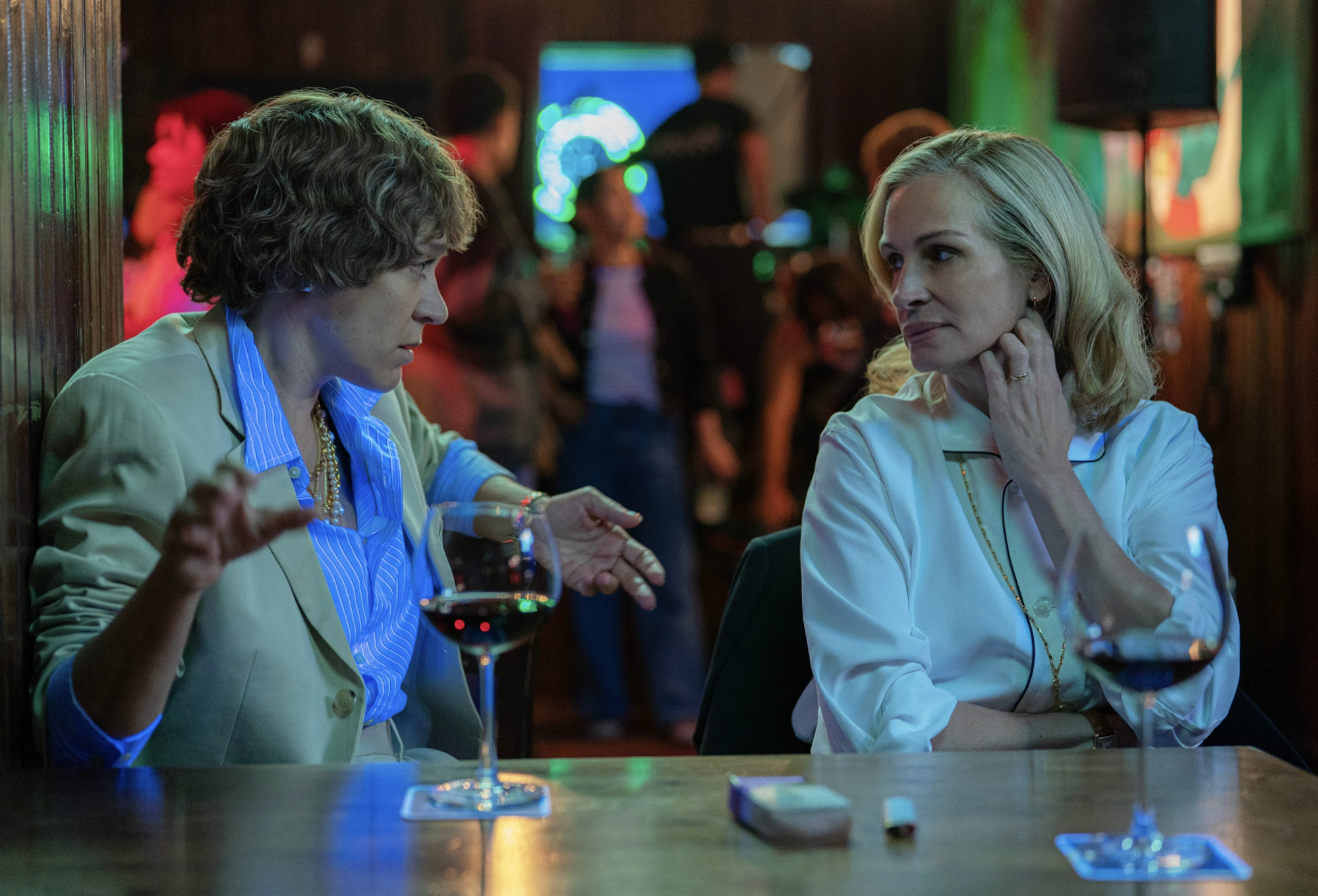 Two middle aged white women in smart office attire sit in a wood-paneled bar under neon lighting. They are discussing something.