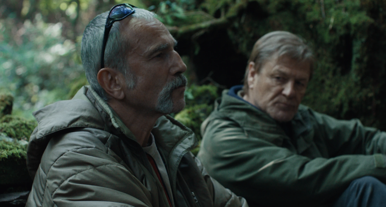 Two older white men sit in a wooded area wearing waterproof winter coats and joyless expressions.