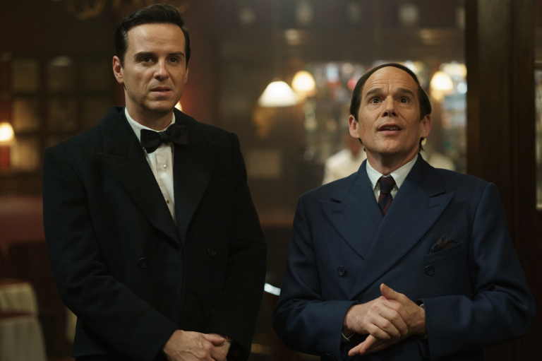 A handsome middle aged white man in a tuxedo stands next to a goofier-looking man in a blue suit and tie who is clasping his hands together gleefully.