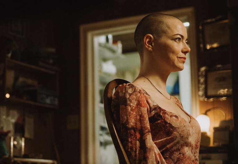 A woman with a shaved head, wearing a traditional dress, stands in side profile in the room of a dimly lit house.