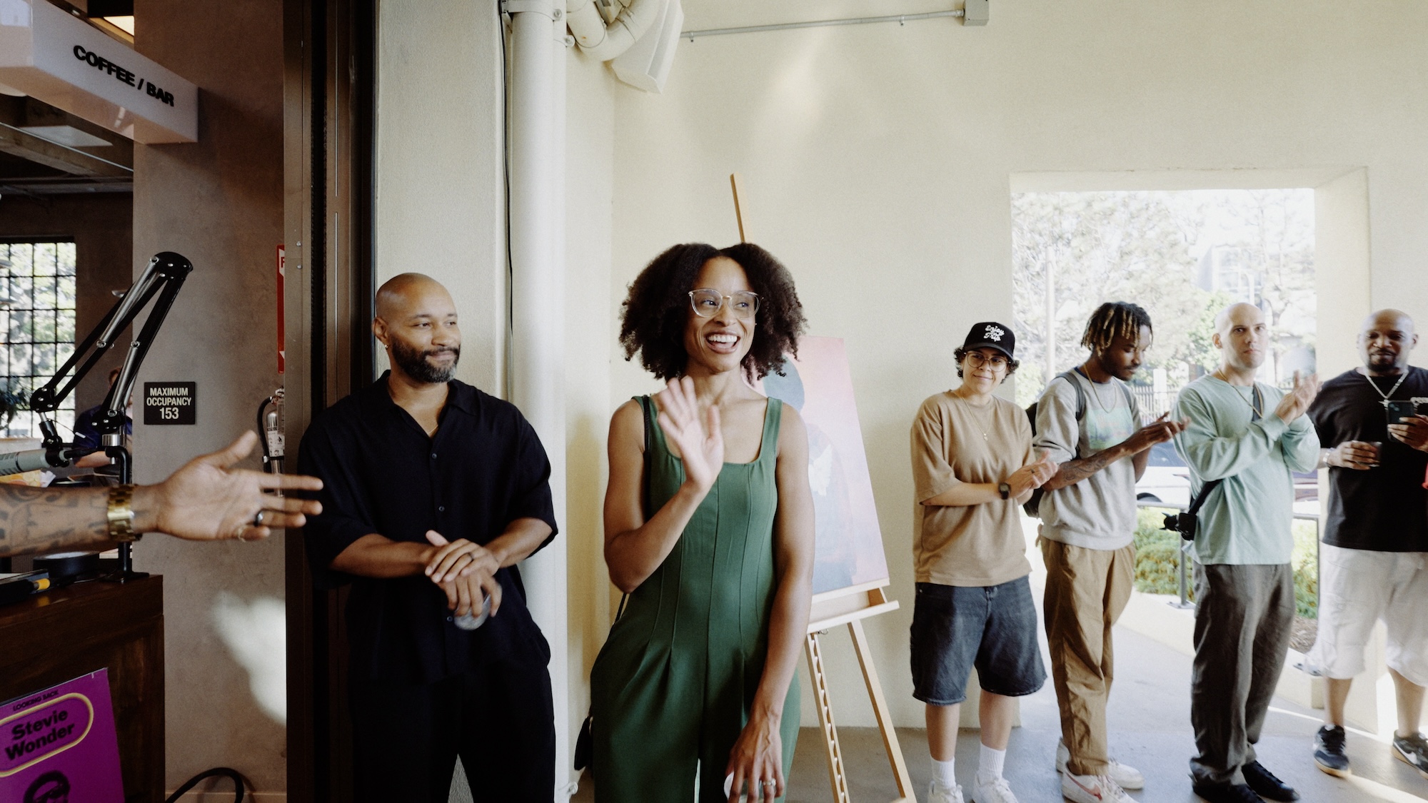 A brown skin woman in a green jumper and glasses waves to a crowd of people in an art studio space. 