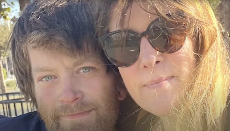 Close up photo of a white woman with sunglasses on, next to a younger white man with a scruffy beard. Their heads are positioned side by side and close together.