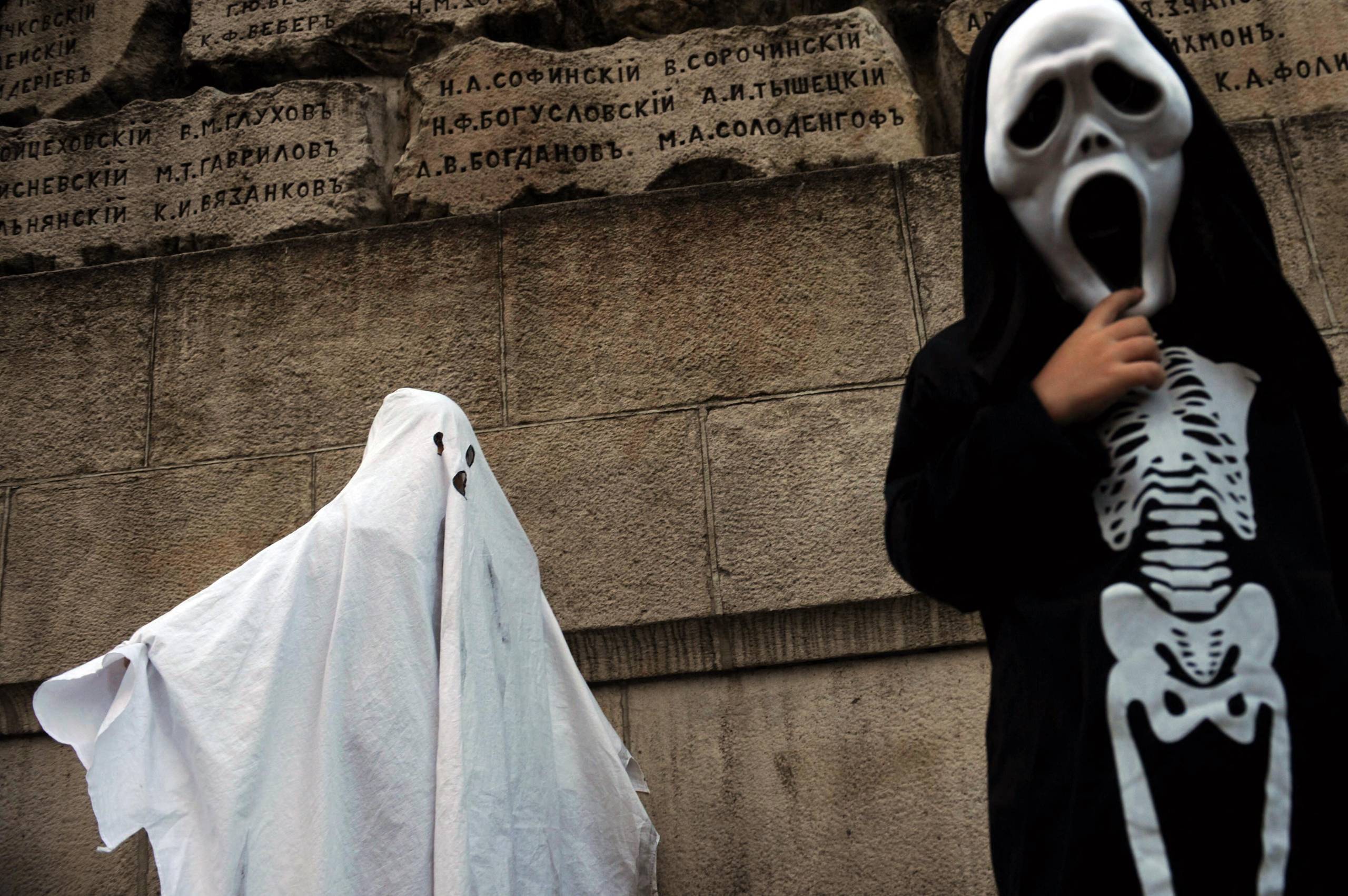 A photo of a child on the left, draped in a white sheet as part of a ghost costume. A child on the right is dressed in a skeleton costume and a 'Scream' mask.