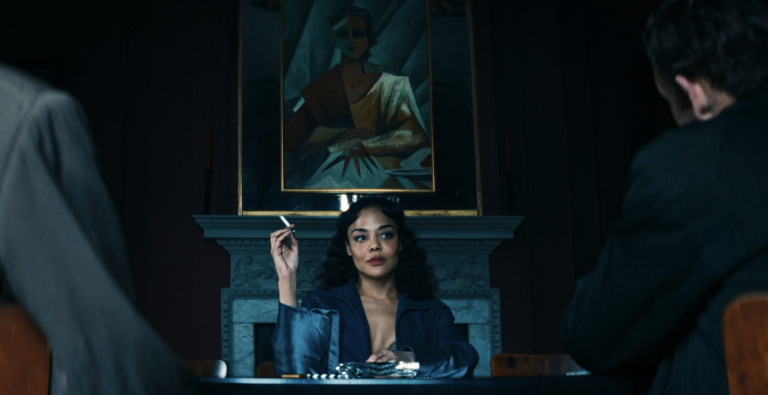 A beautiful mixed race woman sits behind a grand desk in a darkened office, a classic painting hanging behind her above a marble fireplace.