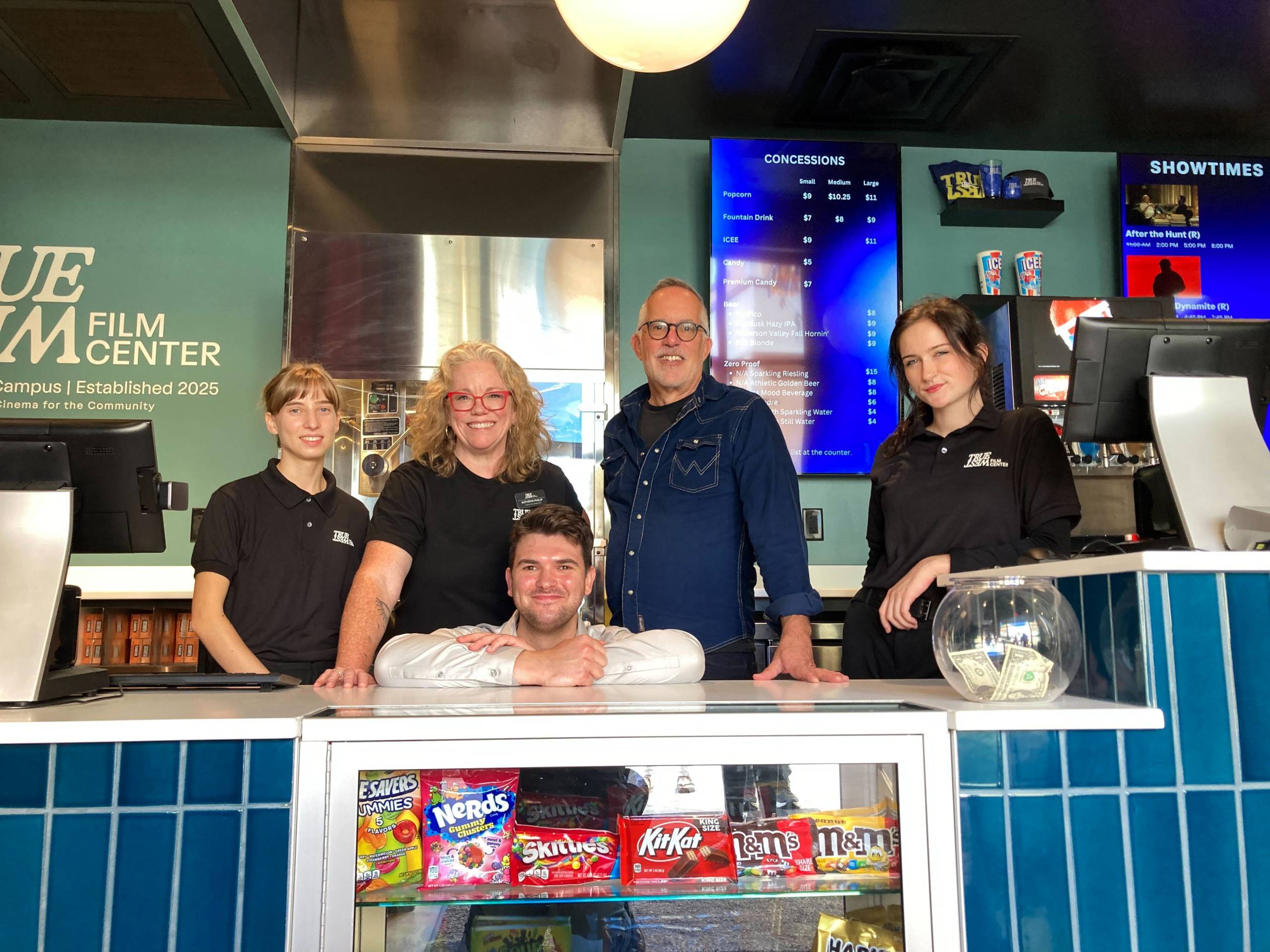 Five white people of varying ages gather to pose behind a blue snack counter displaying boxes of candy in the front and prices for popcorn and drinks in the back