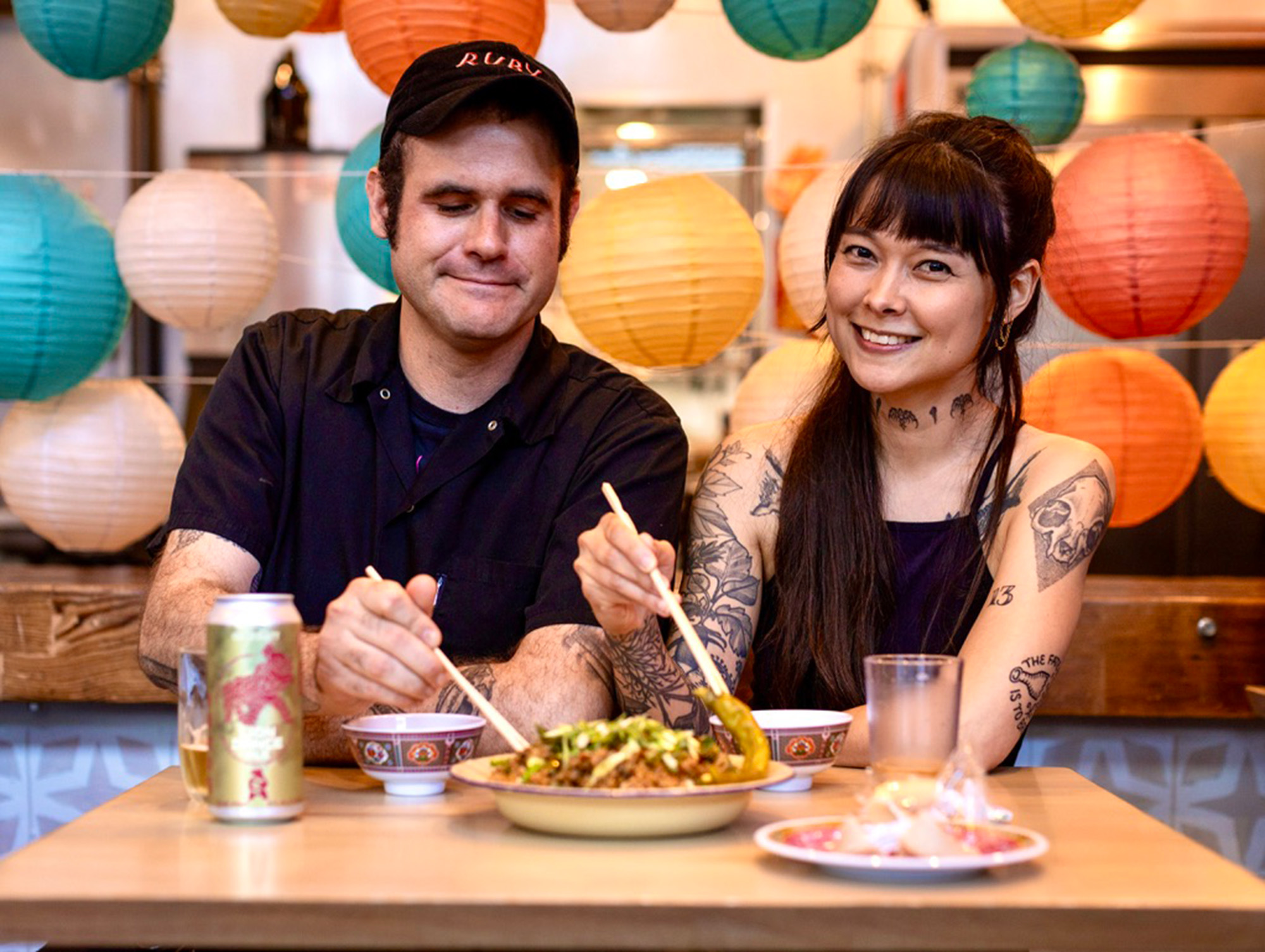 A couple holding chopsticks to pick at a plate of food poses for a portrait.