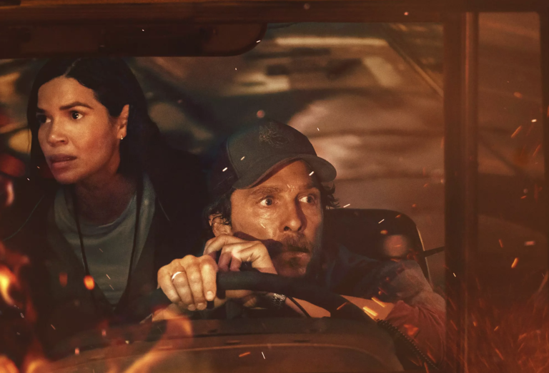 A stressed-looking Latina peers over the shoulder of a scared looking white male bus driver who is seated at the wheel. Fire reflects on the windshield.