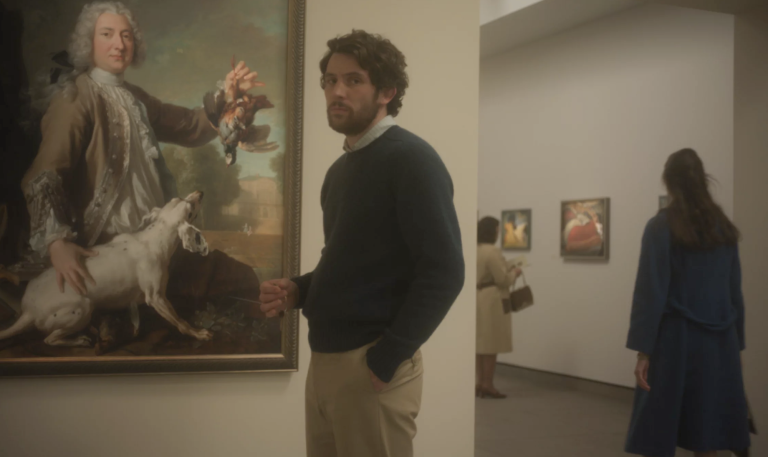 A white man in his 30s stands sheepishly in an art museum. He is wearing a shirt, sweater and slacks.