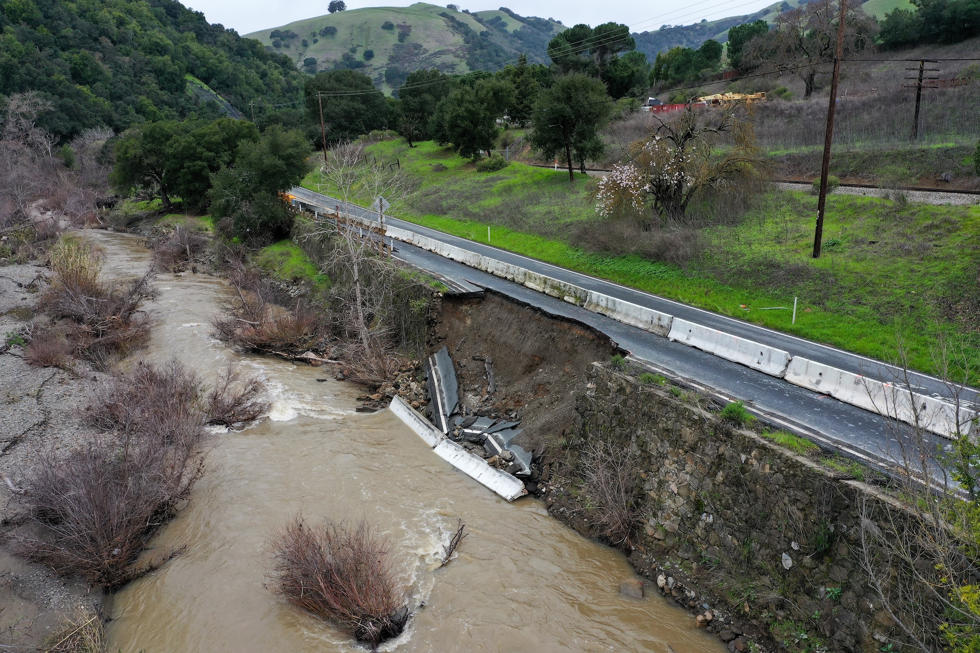 A narrow road surrounded by stark hills, running past a creek full of brown water, has a chunk missing. The missing section of the road can be seen near the water below.
