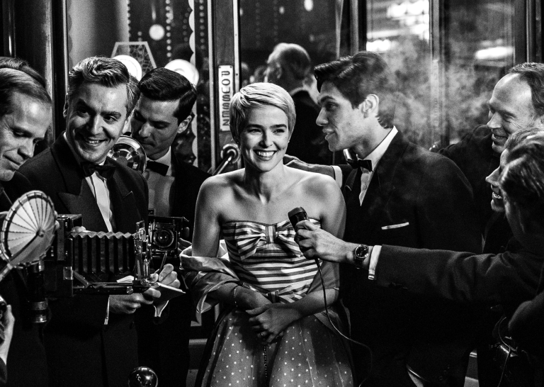 A young beautiful white woman with cropped blond hair wearing a strapless gown, stands smiling, surrounded by reporters holding microphones to her face.