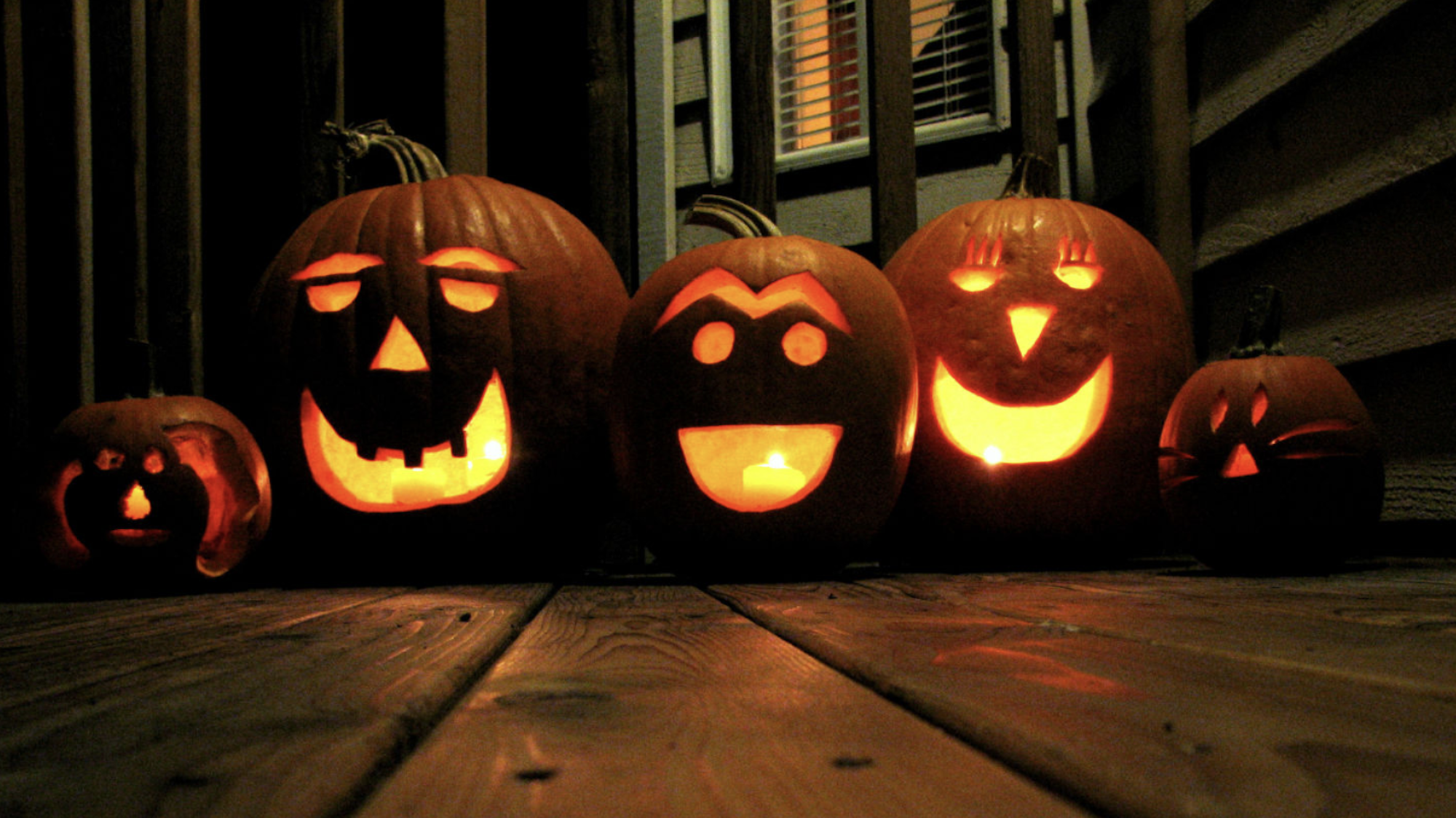 Three large, carved jack-o-lanterns sit lit up on a porch, with two smaller versions either side.