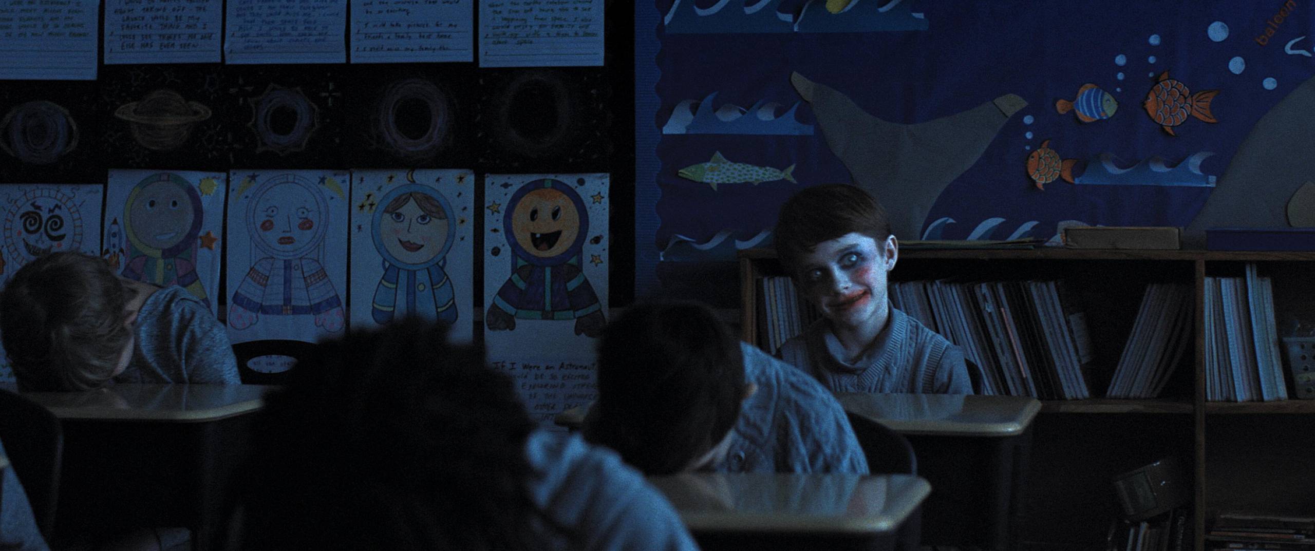 An eerie, smiling child wearing facepaint sits, head cocked to one side, at the back of a gloomy, dimly lit classroom. The classmates around him all sit with their heads on the tables in front of them.