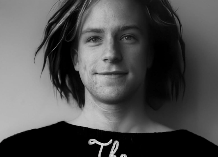 A black and white image of a young white man with chin-length dreadlocks standing against a blank wall and smiling sweetly. He is wearing a black sweater.