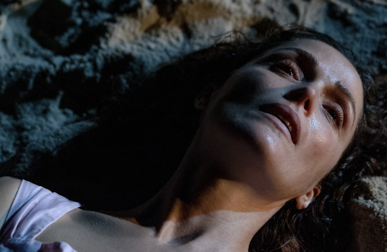 A woman's face in close-up as she lies in the sand. Her face is damp with sweat, her expression entirely blank.