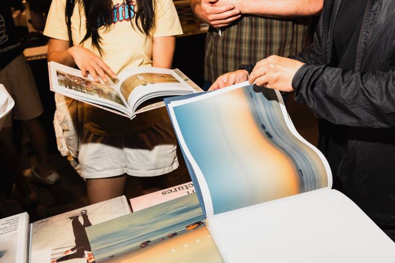 Two people thumb through large photography books next to a book-covered table. A bystander observes.