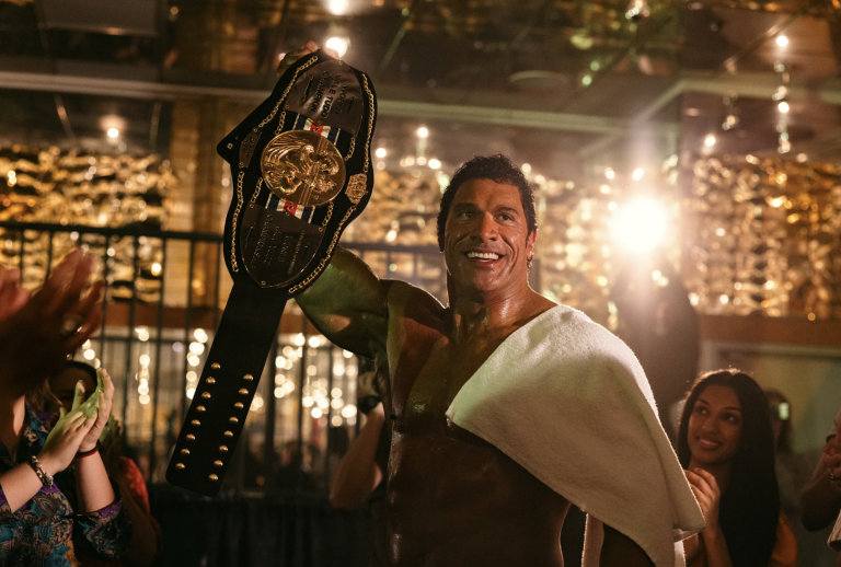 A large, very muscular man stands in the middle of applauding crowds waving a wrestling title belt in the air. He is smiling broadly.