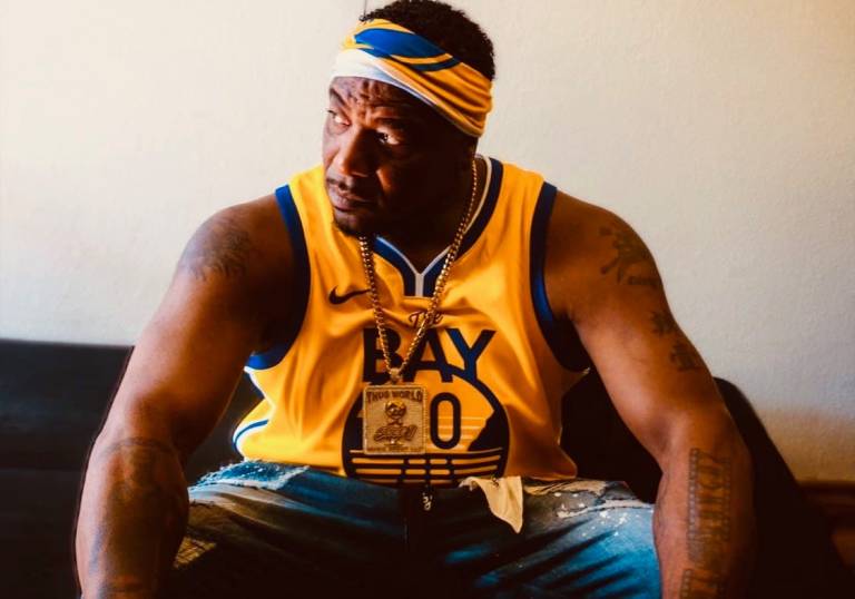 A Black man in a yellow printed tank top and headband sits looking off to the left