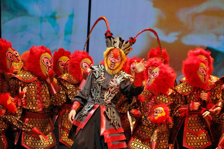 A man in an ornate costume with a feathery headpiece sings in front of a chorus in masks and red-and-gold outfits