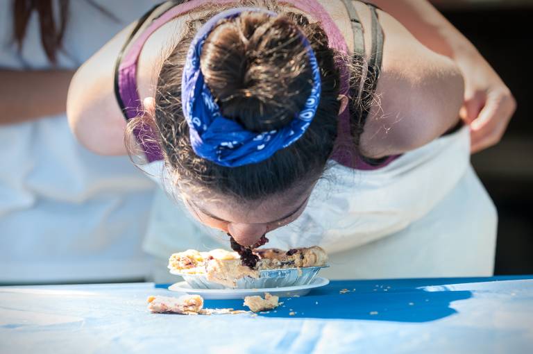 A young girl, with hands behind her back, competes in a blueberry pie eating contest at a country fair.