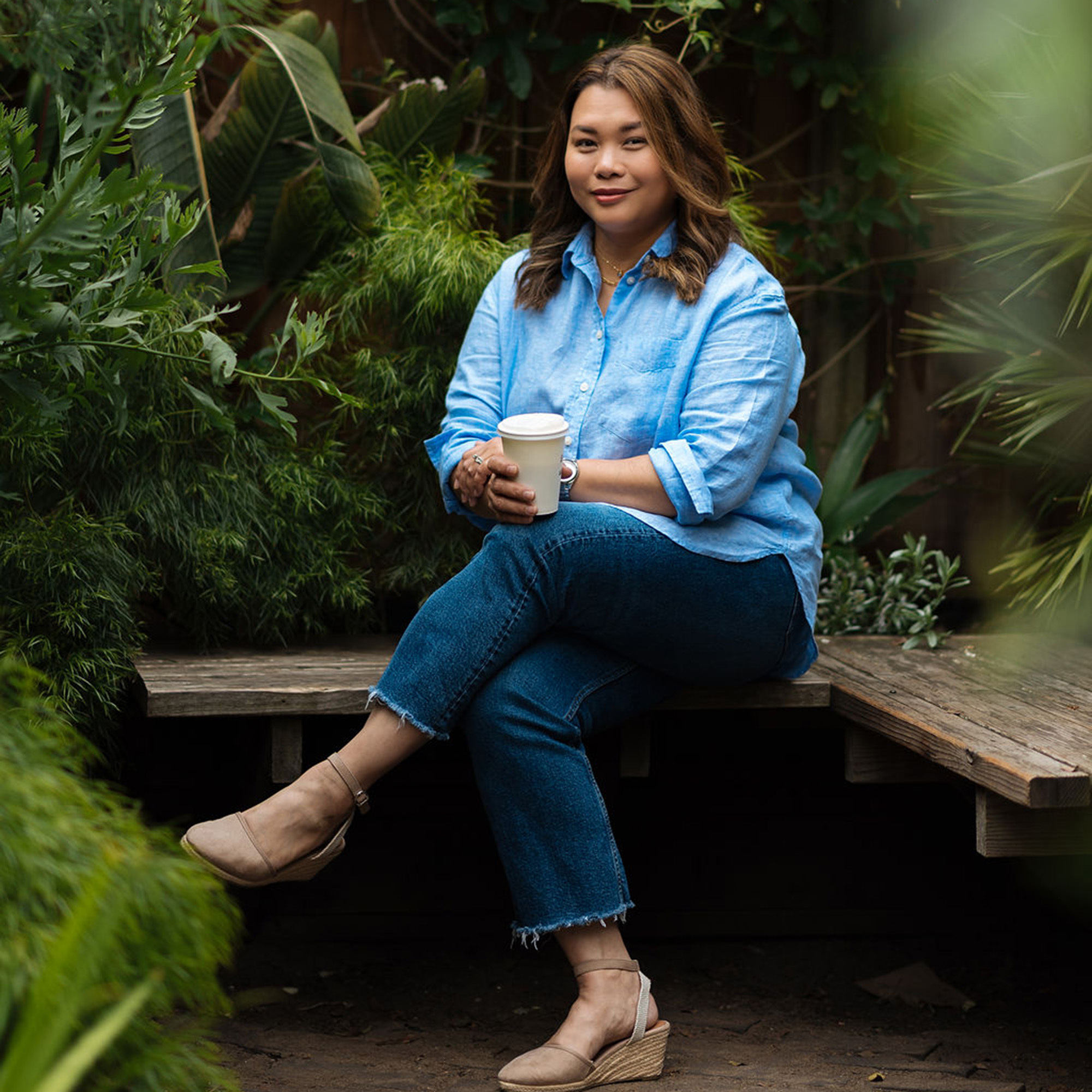 A Filipino woman poses for a portrait while holding a coffee mug, sitting on a wooden bench in some lush green garden setting.