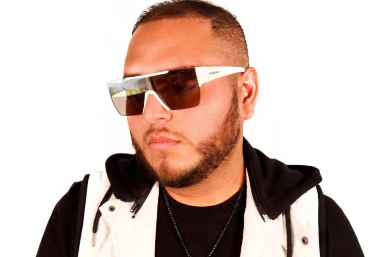 A man in sunglasses, a white vest and black shirt stands with his hands in front of him as he poses for a photo.