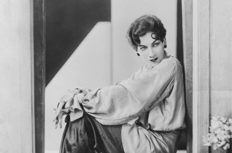 A coiffed and made-up gender nonconforming person sitting elegantly in a doorway, dressed in silky blouse, pants and high heels.