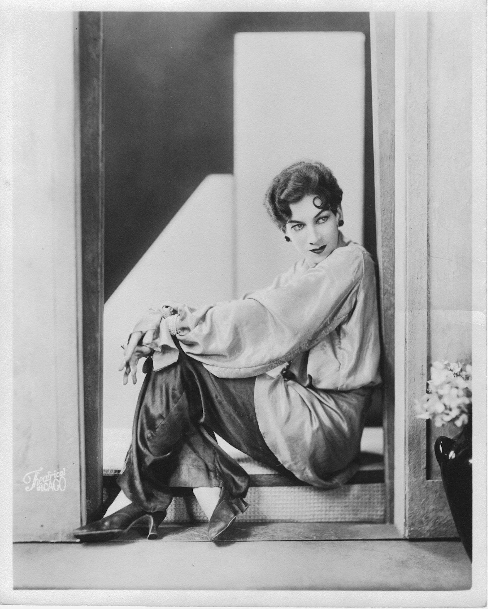 A coiffed and made-up gender nonconforming person sitting elegantly in a doorway, dressed in silky blouse, pants and high heels.